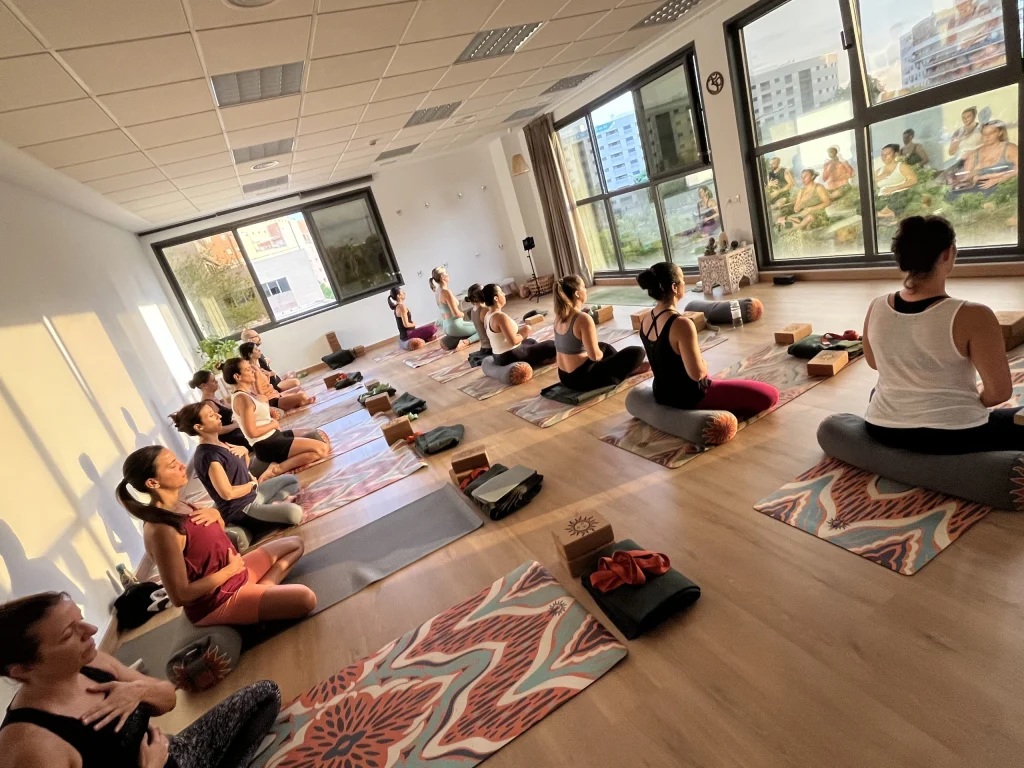 Grupo de alumnas sentadas en sus esterillas durante una clase, en una sala amplia con grandes ventanales